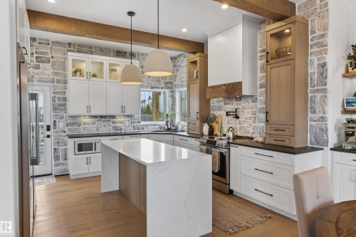 Gourmet kitchen featuring stone accent walls, wood-finish flooring, a central island with a waterfall countertop, white cabinetry with glass display fronts, and a built-in microwave - 711 Beach Avenue, Cold Lake, AB - Indoor Photo Showing Kitchen With Upgraded Kitchen