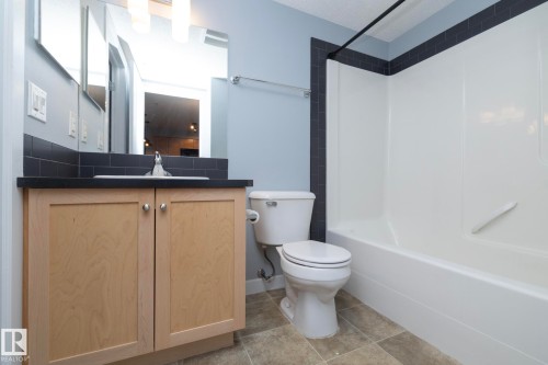 Bathroom featuring a light wood-finish vanity with a dark countertop, an integrated sink, a large mirror, and a white tub with dark subway tile surround - 208 3719 Whitelaw Lane, Edmonton, AB 