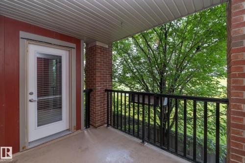 Outdoor balcony featuring brick and wood-paneled walls, an overhead ceiling, a full-lite door with internal blinds, and a black metal railing - 208 3719 Whitelaw Lane, Edmonton, AB 