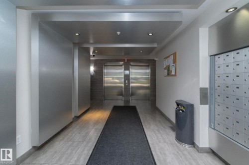 Building entrance hallway featuring light-toned tile flooring, recessed ceiling lighting, two stainless steel elevators, and a wall of individual mailboxes - 208 3719 Whitelaw Lane, Edmonton, AB 