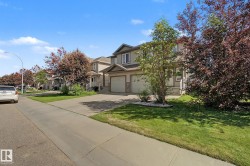 Two-story residence featuring a neutral-toned exterior with stone accents, a two-car garage, established landscaping, a concrete driveway, and a paved sidewalk - 