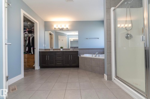 Bathroom featuring a dual-sink vanity with dark wood cabinetry, a built-in bathtub with tiled surround, a glass-enclosed shower, light-toned tile flooring, and a walk-in closet - 2016 Redtail Common, Edmonton, AB 