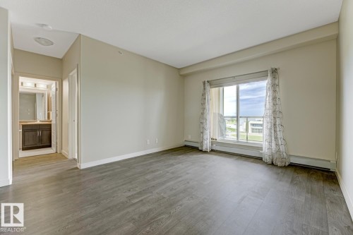 Spacious room featuring wood-finish flooring, a large window, light-colored walls, and white baseboards - 450 11517 Ellerslie Road, Edmonton, AB 