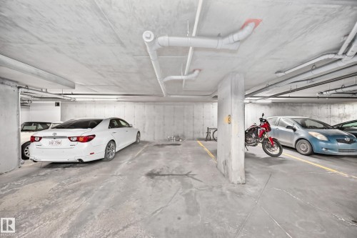 Concrete parking garage with exposed ceiling pipes, fluorescent strip lighting, and painted yellow parking lines - 308 18126 77 Street, Edmonton, AB 