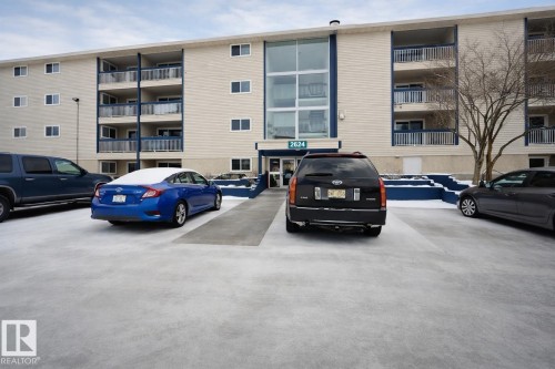 Multi-story building featuring light-colored siding, multiple balconies with dark railings, and a central glass entrance - 107, 2624 Millwoods Road East Nw, Edmonton, AB - Outdoor