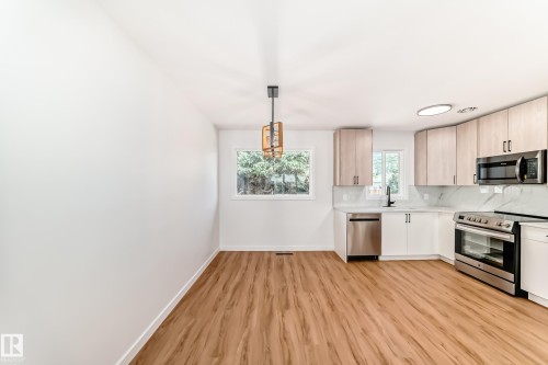 Open-concept kitchen and dining area featuring wood-finish flooring, light wood cabinetry, stainless steel appliances, a white tile backsplash, and a modern ceiling fixture - 4842 116Ave, Edmonton, AB - Indoor Photo Showing Kitchen