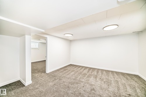 Spacious interior room featuring plush gray carpeting, white painted walls, recessed ceiling lighting, and a visible window opening into an additional space - 4842 116Ave, Edmonton, AB - Indoor Photo Showing Other Room