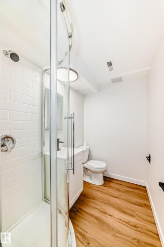 Compact bathroom featuring a curved glass shower enclosure, subway tile shower surround, wood-finish flooring, a white vanity with a rectangular sink, and a modern toilet - 4842 116Ave, Edmonton, AB - Indoor Photo Showing Bathroom