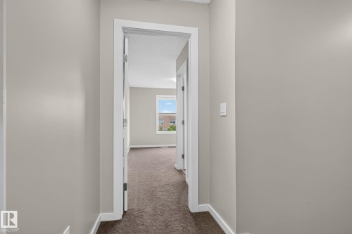 Hallway featuring neutral-toned walls, white trim, and brown carpet leading into a room with a window - 57 3710 Allan Drive, Edmonton, AB 