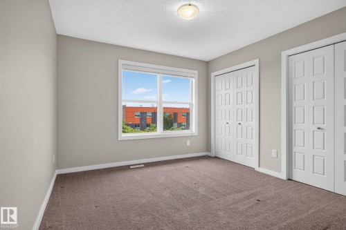 Bedroom featuring a large window, two sets of bi-fold closet doors, neutral wall paint, white trim, and brown carpet flooring - 57 3710 Allan Drive, Edmonton, AB 