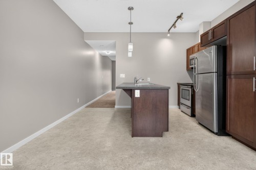 Kitchen featuring rich wood cabinetry, stainless steel appliances, a center island with a sink, pendant lighting, and light-toned flooring - 57 3710 Allan Drive, Edmonton, AB 
