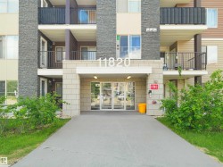 Building exterior featuring a stone facade, multiple balconies with dark railings, a prominent entrance with glass doors, and a concrete pathway - 
