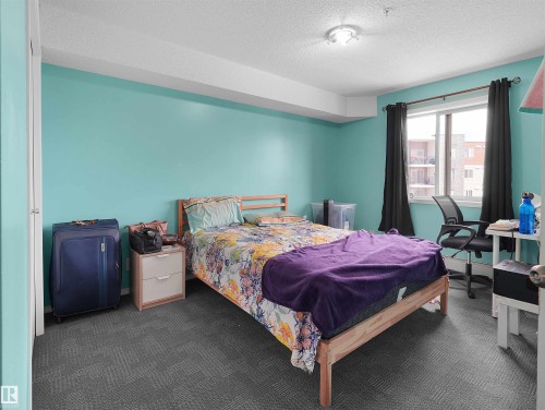 Bedroom featuring a window with black curtains, textured gray carpet, and light blue walls - 313 11820 22 Avenue, Edmonton, AB 