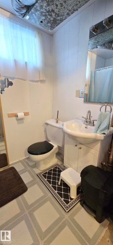 Bathroom featuring light-toned walls, a white sink with a vanity cabinet, a toilet, a mirror, and a window with a sheer curtain - 5304 49 Street, Two Hills, AB - Indoor Photo Showing Bathroom