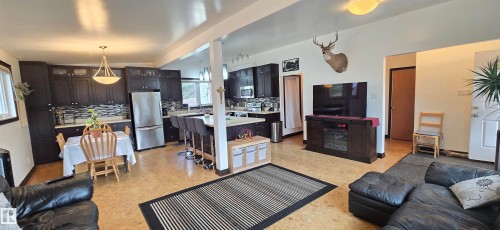 Open-concept living space featuring wood-finish flooring, a contemporary kitchen with dark cabinetry and stainless steel appliances, and a breakfast bar with pendant lighting - 5304 49 Street, Two Hills, AB - Indoor