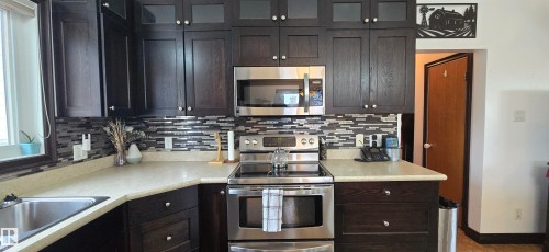 Kitchen featuring dark wood-finish cabinetry with glass-front upper cabinets, light-toned countertops, and a full tile backsplash - 5304 49 Street, Two Hills, AB - Indoor Photo Showing Kitchen