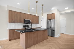 Modern kitchen featuring wood-finish cabinetry, stainless steel appliances, a black island countertop with a sink, white subway tile backsplash, and light wood-finish herringbone flooring - 