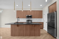 Kitchen featuring a central island with a dark countertop and integrated sink, pendant lighting, wood-finish cabinetry, stainless steel appliances, and herringbone-pattern wood-finish flooring - 