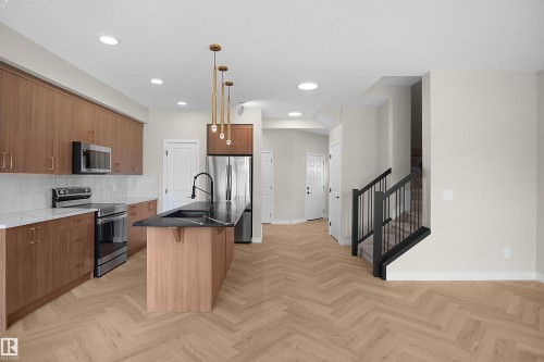 Open-concept kitchen featuring wood-finish cabinetry, stainless steel appliances, a center island with a black countertop and integrated sink, gold-tone pendant lighting, and herringbone wood-finish flooring - 8523 Mayday Run, Edmonton, AB 