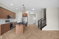 Open-concept kitchen featuring wood-finish cabinetry, stainless steel appliances, a center island with a black countertop and integrated sink, gold-tone pendant lighting, and herringbone wood-finish flooring - 