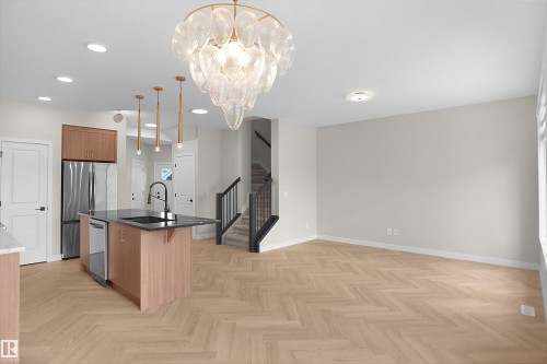 Open concept living space featuring a contemporary chandelier, herringbone wood-finish flooring, a kitchen island with dark countertops, stainless steel appliances, and a modern staircase with dark railings - 8523 Mayday Run, Edmonton, AB 