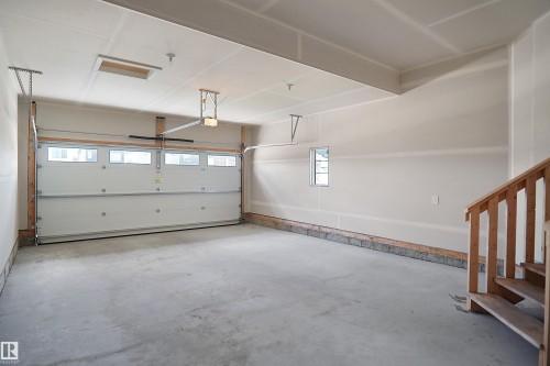 Spacious garage with concrete flooring, a white sectional overhead door with upper window panels, and a wood staircase with railing - 8523 Mayday Run, Edmonton, AB 