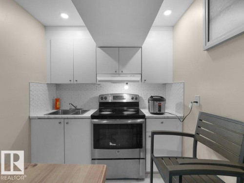 Compact kitchen area featuring a stainless steel range, white cabinetry, a double basin sink, light-toned countertops, and a geometric-patterned backsplash - 1419 27 Av, Edmonton, AB 