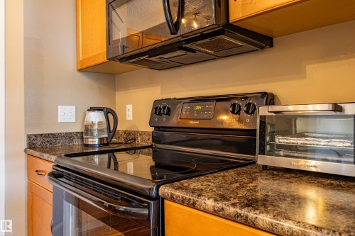 Kitchen featuring a black electric range with an overhead microwave, dark speckled countertops, and light wood cabinetry - 13 1816 Rutherford Road, Edmonton, AB 