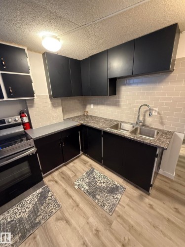 Contemporary kitchen featuring black cabinetry, light wood-finish flooring, a stainless steel double sink, and a light-toned subway tile backsplash - 10220 107 Street, Westlock, AB - Indoor Photo Showing Kitchen With Double Sink