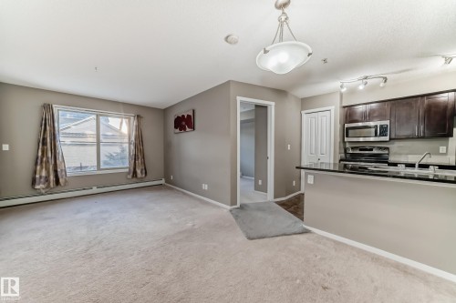 Open concept living area featuring carpeted flooring and an integrated kitchen with dark wood-finish cabinetry, stainless steel appliances, and a black countertop - 121 111 Watt Common, Edmonton, AB 