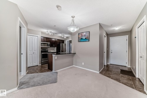Open-concept living space featuring a kitchen with dark wood-finish cabinetry, stainless steel appliances, and a black countertop with a breakfast bar - 121 111 Watt Common, Edmonton, AB 