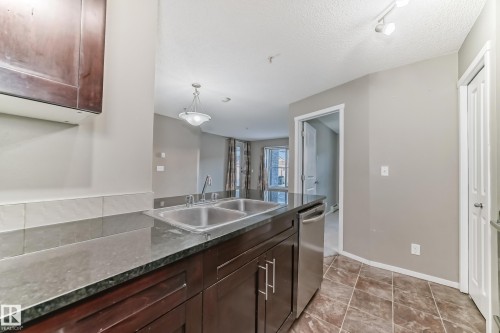 Kitchen featuring a double basin stainless steel sink, dark wood-finish cabinetry with silver hardware, stone countertops, and a full-size stainless steel dishwasher - 121 111 Watt Common, Edmonton, AB 