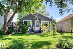 Gray stucco exterior featuring a white front door, white window trim, well-maintained front lawn, mature tree, and planted garden beds - 
