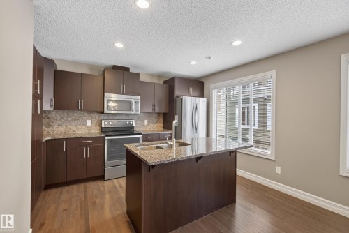 Modern kitchen featuring wood-finish flooring, dark wood cabinetry, stainless steel appliances, a tile backsplash, and an island with a stone-look countertop and integrated sink - 53 3625 144 Avenue, Edmonton, AB 