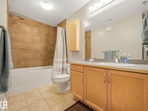 Full bathroom featuring a tub and shower combination with brown wall tile surround, a single vanity with light wood cabinetry, a light-colored countertop, and a large mirror - 150 4827 104A Street, Edmonton, AB 