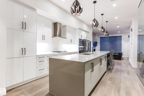 Sleek kitchen featuring high-gloss white cabinetry, a large central island with an integrated sink, stainless steel appliances, and wood-finish flooring - 13218 102 Avenue, Edmonton, AB 
