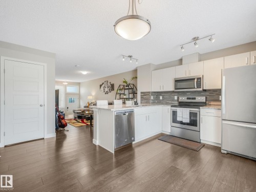 Open-concept kitchen featuring wood-finish flooring, white cabinetry, stainless steel appliances, a stone countertop, and a tiled backsplash - 55 4470 Prowse Road, Edmonton, AB 