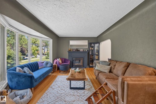 Living space featuring a prominent bay window, a central fireplace with a dark mantel, wood-finish flooring, and a textured ceiling - 11015 122 Street, Edmonton, AB 