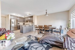 Open-concept living space featuring wood-finish flooring, a kitchen with dark cabinetry and stainless steel appliances, a kitchen island with seating, a dining area with overhead lighting, and a window with blinds - 
