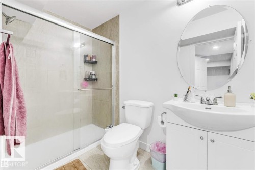 Full bathroom featuring a shower-tub combination with sliding glass doors, neutral-toned shower surround tiling, a white vanity with cabinet storage, a curved basin sink, and a circular wall mirror - 1715 27 Street, Edmonton, AB 