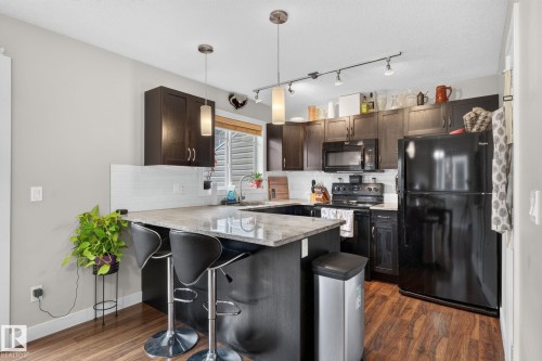 Kitchen featuring dark wood-finish cabinetry, black appliances, a white subway tile backsplash, a U-shaped island with a light-colored countertop, and wood-finish flooring - 89 9535 217 Street, Edmonton, AB 