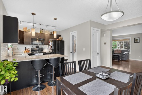 Open-concept kitchen and dining area featuring warm wood-finish flooring, a kitchen island with seating, a full suite of black appliances, and pendant lighting - 89 9535 217 Street, Edmonton, AB 