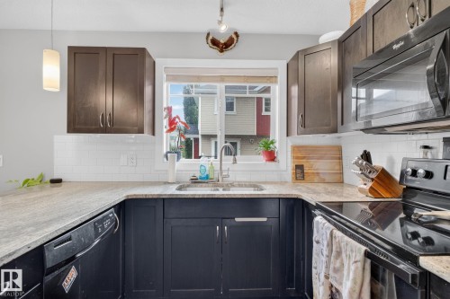 U-shaped kitchen featuring dark wood cabinetry, light-colored countertops, a double basin sink with a window overhead, a built-in microwave, and a black electric range - 89 9535 217 Street, Edmonton, AB 