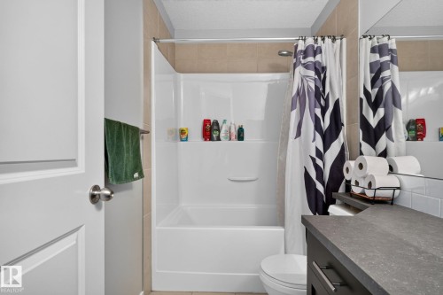 Full bathroom featuring a white tub and shower combination with tan wall tiling, a dark vanity with a textured countertop, and a white toilet - 89 9535 217 Street, Edmonton, AB 