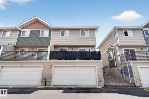 Townhome exterior featuring light-colored horizontal siding, a prominent gable with brown shake siding and green vertical siding, two single-car garages with white doors, and a second-story balcony with black metal railings - 89 9535 217 Street, Edmonton, AB 