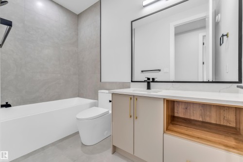 Contemporary bathroom featuring a white soaking tub with gray tiled surround, modern toilet, and a vanity with light cabinetry, wood-finish open shelving, and a white countertop - 2611 62 Avenue, Rural Leduc County, AB - Indoor Photo Showing Bathroom