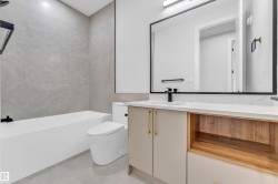 Contemporary bathroom featuring a white soaking tub with gray tiled surround, modern toilet, and a vanity with light cabinetry, wood-finish open shelving, and a white countertop - 