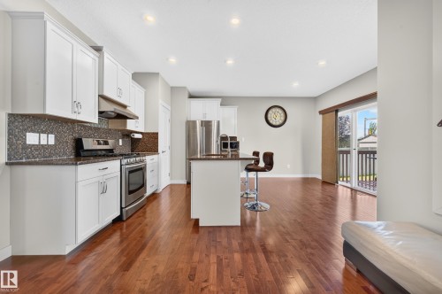 Open-concept kitchen and living area featuring wood-finish flooring, white cabinetry, stainless steel appliances, a kitchen island with seating, and sliding glass doors to an exterior deck - 1512 Cunningham Cape, Edmonton, AB 