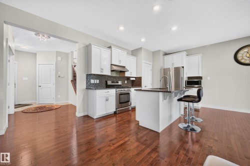 Open-concept kitchen featuring white cabinetry, dark stone countertops, stainless steel appliances, a mosaic tile backsplash, and wood-finish flooring - 1512 Cunningham Cape, Edmonton, AB 