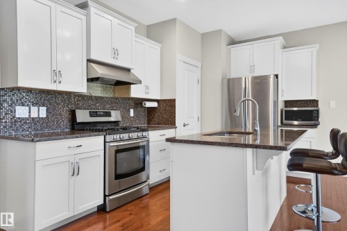 Modern kitchen featuring white cabinetry, stainless steel appliances, a mosaic tile backsplash, and a breakfast bar with stone-finish countertops - 1512 Cunningham Cape, Edmonton, AB 